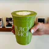 16 oz plastic cup with white "The Juice Shop" logo, green smoothie with coconut, held by manicured hand, white wall and shelf with photos
