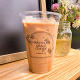 20 oz recyclable plastic cup with black "Marvelous Grace Coffee" logo, filled with iced milk coffee with syrup, on wooden table with chalkboard and flowers in background