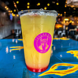 20 oz plastic cup with pink "Yellow King Brews" logo, filled with yellow drink, red syrup, and ice, standing on blue-yellow painted table with pub in background
