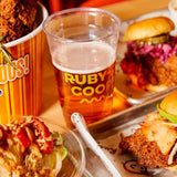 32 oz plastic cup with yellow "Ruby's Coop" logo, filled with beer with froth, surrounded by two burgers and chicken wings in orange bucket