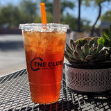 32 oz plastic cup with black "The Club" logo, flat lid and orange straw, filled with iced orange drink, on black mesh table next to succulent pot