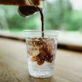 7 oz plastic cup with "Amble Coffee" print and bird logo, espresso shot being poured into sparkling drink, on wooden table by window