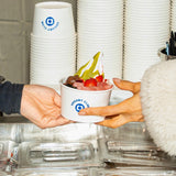 8 oz paper food container with blue "Yogurt Club" logo, filled with ice cream and berries, being passed by two hands in a cafe