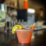 9 oz plastic cup with Social District Downtown text, filled with orange drink with lemon slice and novelty straw, on bar table