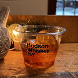9 oz plastic cup with Hudson Whiskey NY text, filled with whiskey and dried orange slice, on wooden table in restaurant