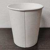 Blank white 10 oz paper cup showing seam, on grey wooden table, white wall in background