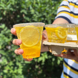 12 oz plastic cup with flat lid, filled with sparkling lemon drink with lemon garnish, held by hand, tray with four more drinks, bush in background
