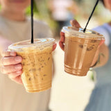 Two 12 oz plastic cups filled with iced milk coffee, with straw lids and black straws, held by people wearing white shirts outdoors