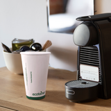 16 oz compostable paper cup with green band and "ecotainer" text, on wooden table beside coffee machine with coffee pods