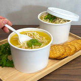 Two 16 oz paper food containers, filled with chicken noodle soup, with a flat lid and spoon, on a wooden tray on a table