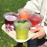 Four 16 oz blank plastic cups with flat lids, filled with colorful slushy drinks, in cardboard carrier held by person in white cardigan, outdoors