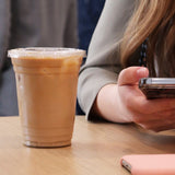 16 oz blank plastic cup with flat lid, filled with iced milk coffee, on wooden table next to person using phone