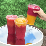 Three 20 oz plastic cups with flat, dome and sip lids, red and yellow smoothies, two on mirror on ground, one lifted by hand, grass in background