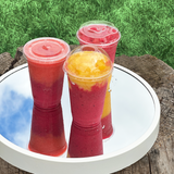 Three 20 oz plastic cups with flat, dome, and sip lids, red and yellow smoothies, standing on mirror on wooden stump, grass in background