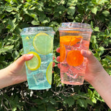 Two 24 oz plastic cups with sip lids, left with iced blue drink and lemon slices, right with clear iced drink and orange slices, held by two hands, bush in background