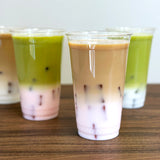 Four 24 oz plastic cups filled with iced coffee with milk and iced matcha with milk, on wooden table, white wall in background