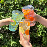 Three 24 oz plastic cups with dome lids, filled with clear drinks with ice and lemon and orange slices, held by three hands, bush in background