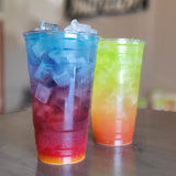 Two 32 oz plastic cups, filled with colorful drinks and ice, on wooden table, cafe in background