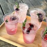 Six 7 oz plastic cups filled with pink iced drink, garnished with berries and rosemary, on light wooden tray on wooden table