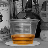 Blank 9 oz recyclable plastic cup filled with orange spirit, on white coaster, with black-and-white alcohol bottles in background