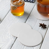 Two blank medium weight round coasters on a white table, next to a honey jar and a tea cup