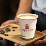 12 oz paper cup with "hoot & sip cafe" pink logo and owl, green band and ecotainer text, on wooden tray with coffee beans, held by person