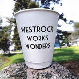 12 oz double-walled paper cup with black "Westrock Works Wonders" text, standing on rock, park in background