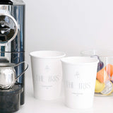 Two 12 oz paper cups with "The Iris" logo and iris graphic, next to coffee machine and glass jar with pods, on white background