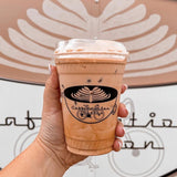 16 oz plastic cup with sip lid and black "Caffeination" logo with coffee graphic, filled with iced milk coffee, held by manicured hand, coffee art wall