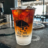20 oz plastic cup with black "R&G Coffee + Racing" text and skeleton logo, iced coffee with milk, on metal gear pattern table, cafe in background