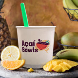 24 oz paper food container, with "Acai Bowls" logo and bowl illustration, with a green spoon, on a table with surrounded by fruits