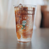 24 oz plastic cup with "Cold is Best" print and coffee character, filled with iced coffee, standing on grey table