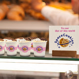 Several 4 oz paper cups with pink "Space Cookies" band and cookie print, filled with whipped dessert and biscuit, on glass display, bakery in background