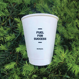 8 oz double-walled paper cup with black "Fuel for Success" logo, leaning on green pine background