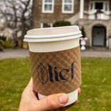 White, paper cups with Kraft Brown Coffee Sleeve displaying "Lief Cafe" logo, posed up by a hand in front of a house