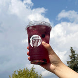 20 oz plastic cup with white "Your Logo Here" print and sip lid, filled with black currant juice, berries and ice, held by hand with blue sky