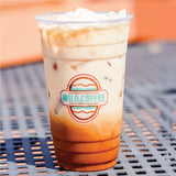 24 oz plastic cup with "Wild Coffee" logo, filled with iced coffee and milk, on blue grid table with orange background