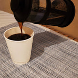 12 oz double-walled paper cup being filled with coffee from a coffee pot, standing on patterned cloth on a table