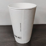Blank white 16 oz premium double walled paper cup showing seam, on grey wooden table, white wall in background