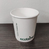 4 oz compostable paper cup with green "ecotainer" text, on grey wooden table, white wall background