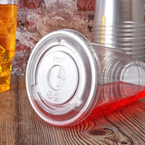 Flat plastic straw lid on 32 oz recyclable plastic cup lying on its side with red liquid, on wooden table, brick wall and other cups in background